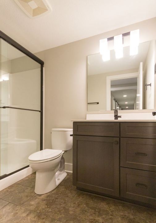 Basement bathroom with mottled brown stone-look tile floor, dark vanity and glass shower enclosure in Avon, IN