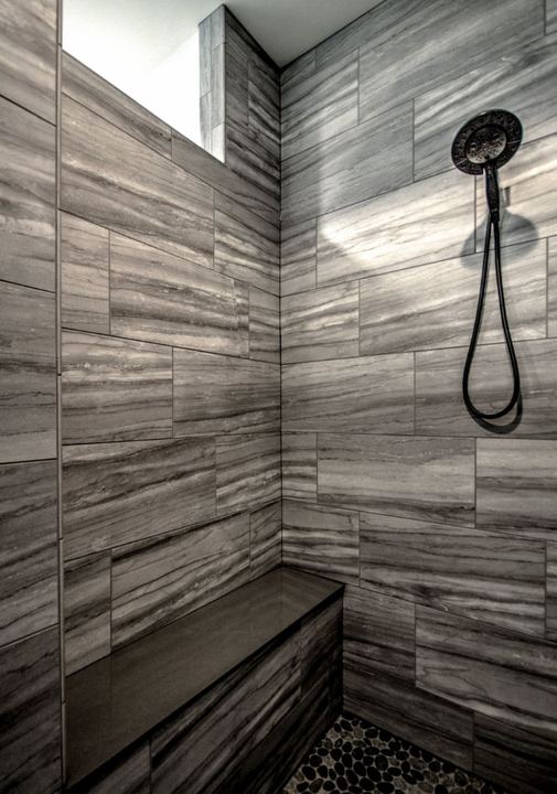 Walk-in shower with gray linear stone-look tile, tiled bench and black pebble floor in Avon, IN