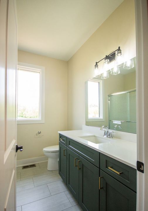 Hall bathroom with gray plank-look tile floor, green shaker vanity, brass pulls and white quartz top in Avon, IN