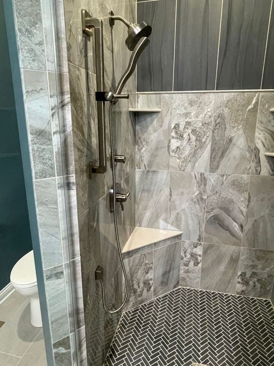 Master shower with gray marble-look tile walls, corner bench, herringbone mosaic floor and slide-bar handheld in Fishers, IN